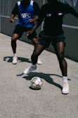 Two people playing street soccer on a concrete surface. One person is wearing a blue jersey with a white logo, while the other is in a black jersey. Both are wearing athletic shoes, and the focus is on the ball at their feet, highlighting movement and agility.