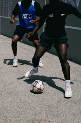 Two people playing street soccer on a concrete surface. One person is wearing a blue jersey with a white logo, while the other is in a black jersey. Both are wearing athletic shoes, and the focus is on the ball at their feet, highlighting movement and agility.