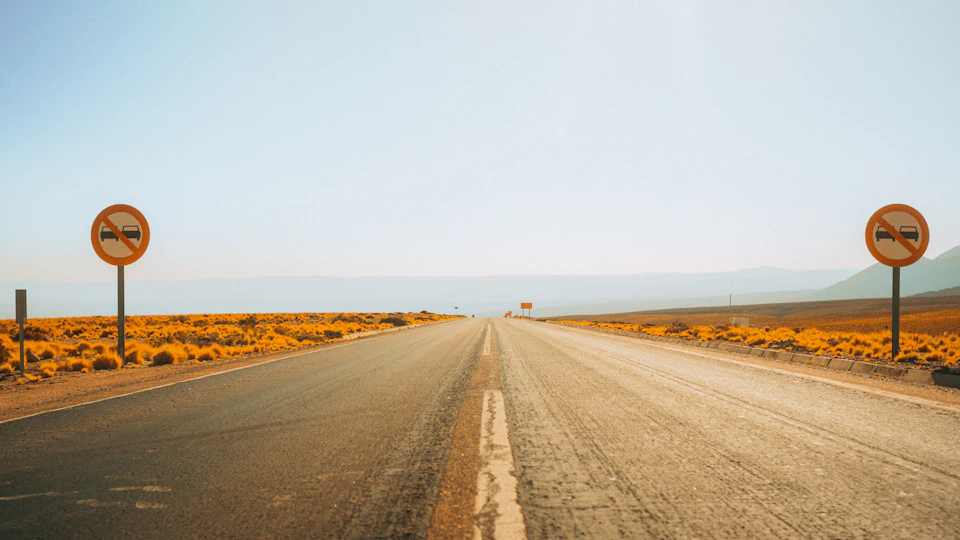 The image depicts a desert road with two no entry signs, one on each side, standing tall against an expansive sky. The road is empty and stretches out into the distance, leading towards a mountain range that forms the backdrop. The scene conveys a sense of solitude and vastness, with the road appearing to be the only human presence in this remote location.