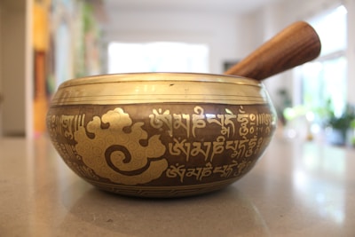 A brass singing bowl with intricate engravings and patterns, including script that appears to be in a language such as Sanskrit or Tibetan. The bowl is placed on a smooth surface, with a wooden mallet beside it. A softly lit room with blurred background elements can be seen, suggesting a peaceful and serene environment.