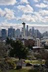 The image depicts a cityscape with a prominent tower resembling the Space Needle in the foreground. The skyline is filled with modern skyscrapers, and a few clouds are scattered across a blue sky. A residential area with trees and smaller buildings is visible in the lower portion of the scene.