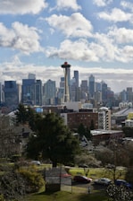 The image depicts a cityscape with a prominent tower resembling the Space Needle in the foreground. The skyline is filled with modern skyscrapers, and a few clouds are scattered across a blue sky. A residential area with trees and smaller buildings is visible in the lower portion of the scene.