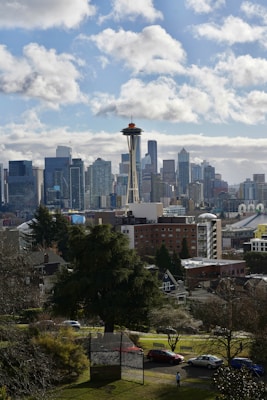 The image depicts a cityscape with a prominent tower resembling the Space Needle in the foreground. The skyline is filled with modern skyscrapers, and a few clouds are scattered across a blue sky. A residential area with trees and smaller buildings is visible in the lower portion of the scene.