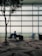 A person sits in an airport terminal next to a large travel bag, with a modern patterned carpet and a silhouetted tree nearby. The seating area is well-lit by natural light through large glass windows, offering a view of an airfield in the distance.