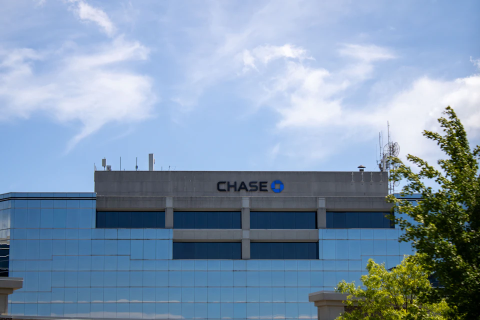 The image shows a large, modern glass and concrete building with a prominent Chase logo on its side. The building is situated under a clear blue sky dotted with fluffy white clouds. In front of the building, there are trees providing a touch of nature to the urban landscape.