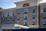 A multi-story hotel building with brick and beige stone exterior featuring numerous windows. Red signage at the top reads 'Hampton Inn & Suites'. The foreground shows part of a vehicle, likely a bus or van.