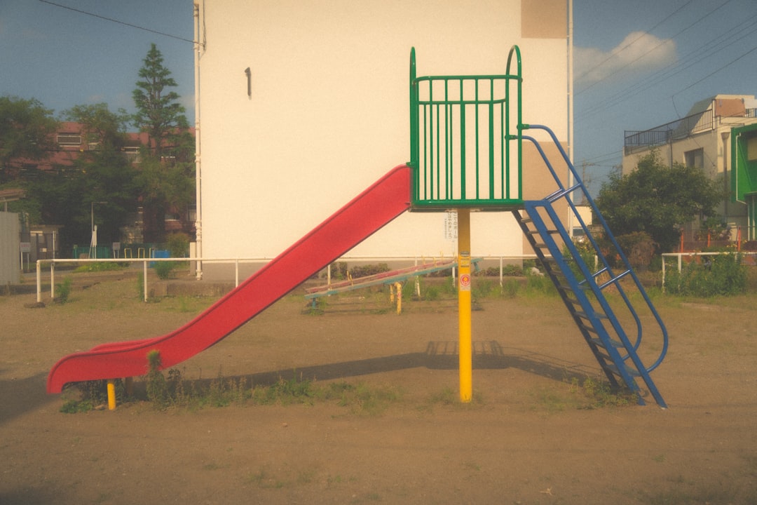 a red slide next to a white building, A small slide in the park. (This photo is Retro style)
