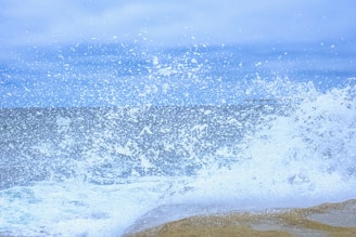 A dynamic scene of ocean waves crashing against the shore, creating a dramatic splash of water droplets against a cloudy sky backdrop.