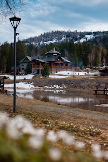 A rustic lodge surrounded by a forest with a snowy mountain backdrop. The scene includes a calm body of water in the foreground and a dirt path leading to the building. The sky is overcast with scattered clouds, adding a serene and peaceful atmosphere.