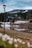 A rustic lodge surrounded by a forest with a snowy mountain backdrop. The scene includes a calm body of water in the foreground and a dirt path leading to the building. The sky is overcast with scattered clouds, adding a serene and peaceful atmosphere.