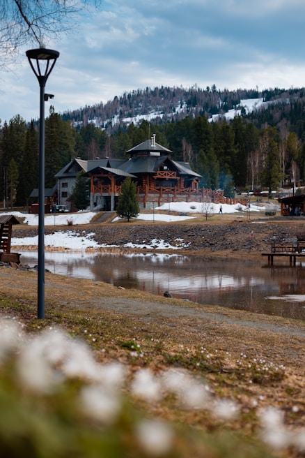 A rustic lodge surrounded by a forest with a snowy mountain backdrop. The scene includes a calm body of water in the foreground and a dirt path leading to the building. The sky is overcast with scattered clouds, adding a serene and peaceful atmosphere.