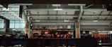 A food court area within an airport terminal features various eateries, including 'Jazz', with illuminated signs and logos. People are walking and seated, and the interior architecture displays modern design elements with skylights and metal framing.