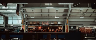A food court area within an airport terminal features various eateries, including 'Jazz', with illuminated signs and logos. People are walking and seated, and the interior architecture displays modern design elements with skylights and metal framing.