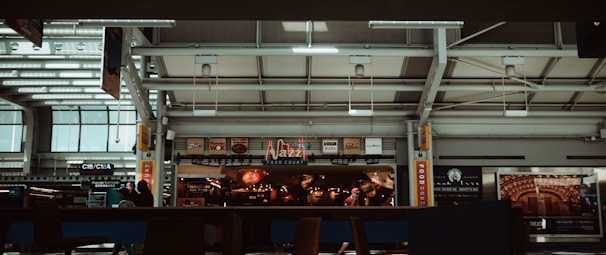 A food court area within an airport terminal features various eateries, including 'Jazz', with illuminated signs and logos. People are walking and seated, and the interior architecture displays modern design elements with skylights and metal framing.