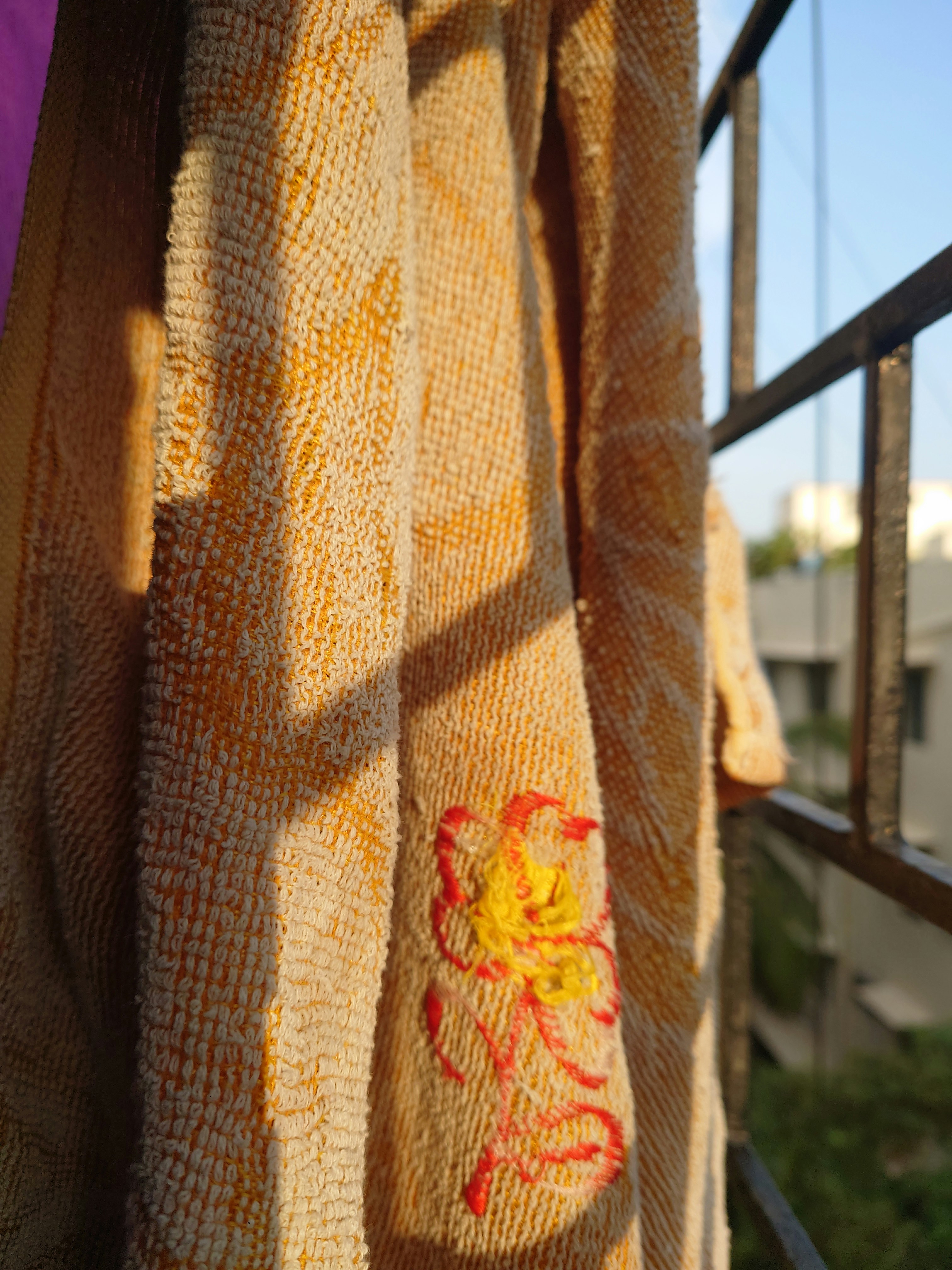 A textured fabric with an embroidered pattern of red and yellow thread hangs from a metal railing. The sunlight casts shadows on the fabric, enhancing the details and textures. The railing is partially in view, with the blurred background of a building and trees.