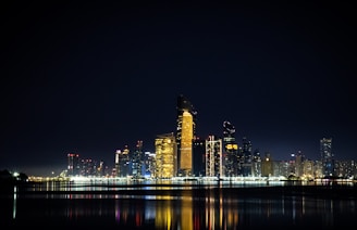A sleek nighttime cityscape with glowing skyscrapers reflecting on a river, bathed in warm golden lights.