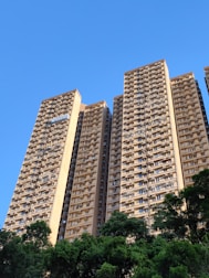 A cluster of tall residential apartment buildings with numerous balconies and windows. The structures are set against a clear blue sky, with lush green trees at the base, adding a natural contrast to the urban architecture.