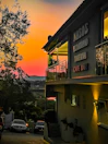 A taverna with signs in Greek stands illuminated under a vibrant sunset sky, featuring hues of orange, pink, and purple. The building has a balcony with glass railing and visible plants. Below, several cars are parked along a narrow road lined with trees, leading towards a scenic landscape.