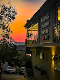 A taverna with signs in Greek stands illuminated under a vibrant sunset sky, featuring hues of orange, pink, and purple. The building has a balcony with glass railing and visible plants. Below, several cars are parked along a narrow road lined with trees, leading towards a scenic landscape.