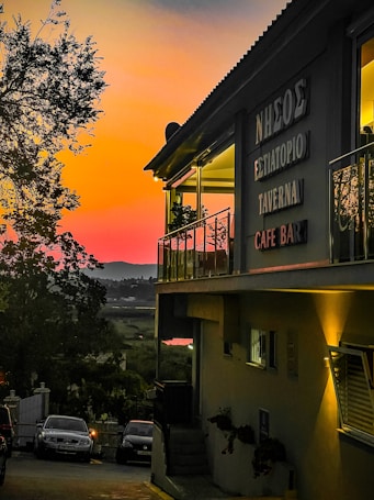 A taverna with signs in Greek stands illuminated under a vibrant sunset sky, featuring hues of orange, pink, and purple. The building has a balcony with glass railing and visible plants. Below, several cars are parked along a narrow road lined with trees, leading towards a scenic landscape.