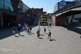 A spacious urban area with people walking and interacting on a large paved courtyard. The scene includes two sets of stairs in the background, lined with green plants and leading up to additional levels filled with more pedestrians. Modern office or commercial buildings of glass and brick surround the area, with natural light illuminating the scene.