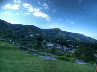A lush green landscape is highlighted with rolling hills under a bright blue sky. A few scattered clouds add contrast to the sky. The foreground features manicured grass and a variety of bushes and plants, while the middle ground showcases a number of buildings partially visible through the trees. The terrain is slightly elevated, providing a clear view of the natural scenery.