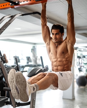 A muscular man is performing a hanging leg raise workout in a gym. He appears focused and is wearing white athletic shorts and socks. The background includes gym equipment such as exercise machines and weights.
