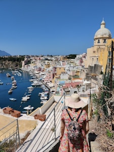 A person wearing a floral dress, straw hat, and backpack walks down a narrow path towards a picturesque coastal town. The town features colorful buildings with a prominent domed building. Several boats are anchored in the calm blue harbor, surrounded by cliffs with greenery.