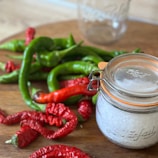 A variety of red and green peppers lie on a wooden surface, with a sealed glass jar containing coarse white salt positioned in the foreground. A clear empty jar can be seen in the background, creating a rustic kitchen scene.