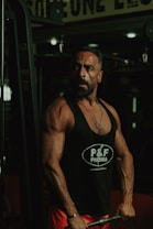 A muscular man is in a gym setting, wearing a black tank top with 'P&F Pharma' printed on it. He appears to be focused and strong, engaged in weightlifting.