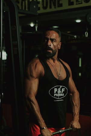 A muscular man is in a gym setting, wearing a black tank top with 'P&F Pharma' printed on it. He appears to be focused and strong, engaged in weightlifting.