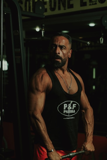 A muscular man is in a gym setting, wearing a black tank top with 'P&F Pharma' printed on it. He appears to be focused and strong, engaged in weightlifting.