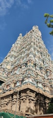 An intricately carved temple tower rises against a clear blue sky, featuring detailed sculptures and ornate decorations in various colors. The architecture displays a traditional South Indian style, characterized by multiple tiers adorned with carvings of deities and geometric patterns. A few green trees partially frame the structure on the right side, adding a natural element to the scene.