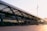 A modern transportation terminal with a sleek architectural design featuring large glass windows and dark metal beams. The structure has two levels, with buses visible on the upper level. There are few people walking and the area is well-lit by sunlight in the late afternoon or early morning.
