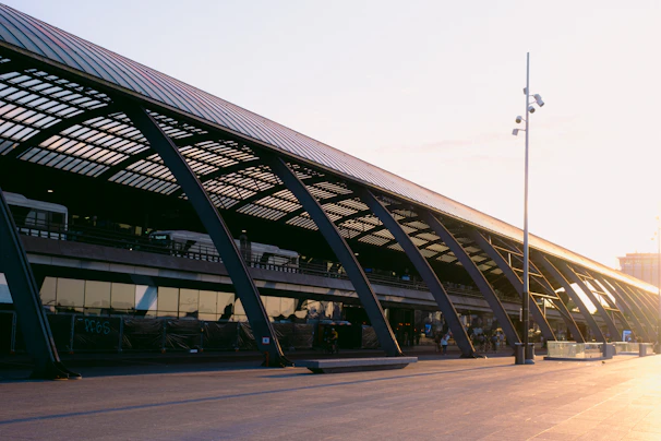 A modern transportation terminal with a sleek architectural design featuring large glass windows and dark metal beams. The structure has two levels, with buses visible on the upper level. There are few people walking and the area is well-lit by sunlight in the late afternoon or early morning.