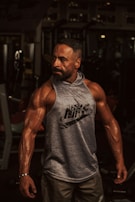 A muscular man stands confidently in a gym environment, showcasing his defined muscles and wearing a sleeveless Nike hoodie. The background is dimly lit, with gym equipment partially visible.