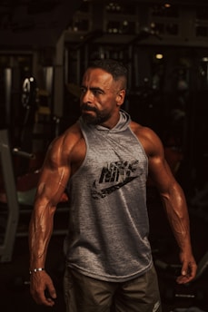 A muscular man stands confidently in a gym environment, showcasing his defined muscles and wearing a sleeveless Nike hoodie. The background is dimly lit, with gym equipment partially visible.