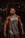 A muscular man stands confidently in a gym environment, showcasing his defined muscles and wearing a sleeveless Nike hoodie. The background is dimly lit, with gym equipment partially visible.