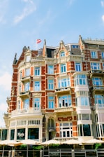 A historic, multi-story building with red brick and light beige stone accents featuring numerous windows and ornate architectural details. A small flag flies on top of the structure. The lower level displays the name 'Marie' and umbrellas can be seen, suggesting a cafe or restaurant setting.