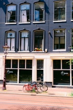 A multi-story building with a dark blue façade adorned with multiple arched windows, some of which have white curtains. Below, there is a restaurant with signage indicating it serves Indonesian cuisine. In front of the building, two bicycles are parked against a metal bike rack on a brick sidewalk. A tall, ornate street lamp is visible on the left side.