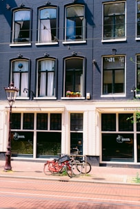 A multi-story building with a dark blue façade adorned with multiple arched windows, some of which have white curtains. Below, there is a restaurant with signage indicating it serves Indonesian cuisine. In front of the building, two bicycles are parked against a metal bike rack on a brick sidewalk. A tall, ornate street lamp is visible on the left side.