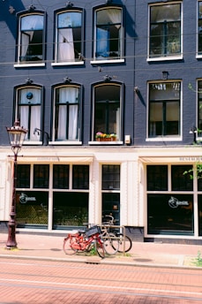 A multi-story building with a dark blue façade adorned with multiple arched windows, some of which have white curtains. Below, there is a restaurant with signage indicating it serves Indonesian cuisine. In front of the building, two bicycles are parked against a metal bike rack on a brick sidewalk. A tall, ornate street lamp is visible on the left side.