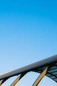 A clear blue sky dominates the image, with the upper segment of a solar panel array visible in the lower part. The structure is supported by sleek metal beams, suggesting a modern, sustainable energy theme.