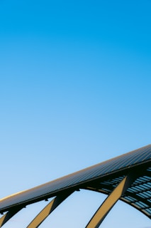 A clear blue sky dominates the image, with the upper segment of a solar panel array visible in the lower part. The structure is supported by sleek metal beams, suggesting a modern, sustainable energy theme.