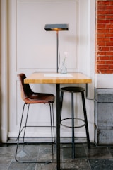 A small wooden table with a metal base is accompanied by a brown leather chair and a black metal stool. A sleek black lamp with a rectangular shade sits on the table, casting light downwards. A single pink flower in a transparent vase adds a touch of elegance. The setting features a mix of white paneling and red brick walls, creating a cozy atmosphere.