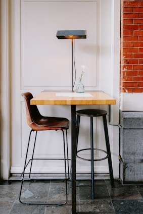 A small wooden table with a metal base is accompanied by a brown leather chair and a black metal stool. A sleek black lamp with a rectangular shade sits on the table, casting light downwards. A single pink flower in a transparent vase adds a touch of elegance. The setting features a mix of white paneling and red brick walls, creating a cozy atmosphere.