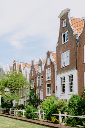 A row of charming, traditional Dutch houses with stepped gables and large windows. The buildings are made of red brick with white detailing. In the foreground, a lush garden with various green plants and flowers is enclosed by a white wooden fence. The sky is clear and bright, giving the scene a serene ambiance.