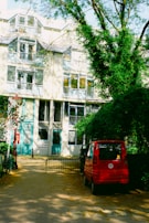 A multi-story residential building features large windows and balconies. In front of the building, there is a paved area with a small red car parked to the side. Green trees with lush leaves partially cover the scene, casting shadows on the pavement and the facade of the building.