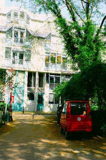 A multi-story residential building features large windows and balconies. In front of the building, there is a paved area with a small red car parked to the side. Green trees with lush leaves partially cover the scene, casting shadows on the pavement and the facade of the building.