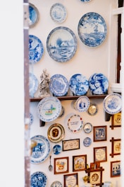 A collection of decorative blue and white ceramic plates is displayed on a wall. The plates feature various designs, including intricate patterns, landscapes, and portraits. Additional items such as framed pictures, a small statue, and other decorative objects complement the plate arrangement.